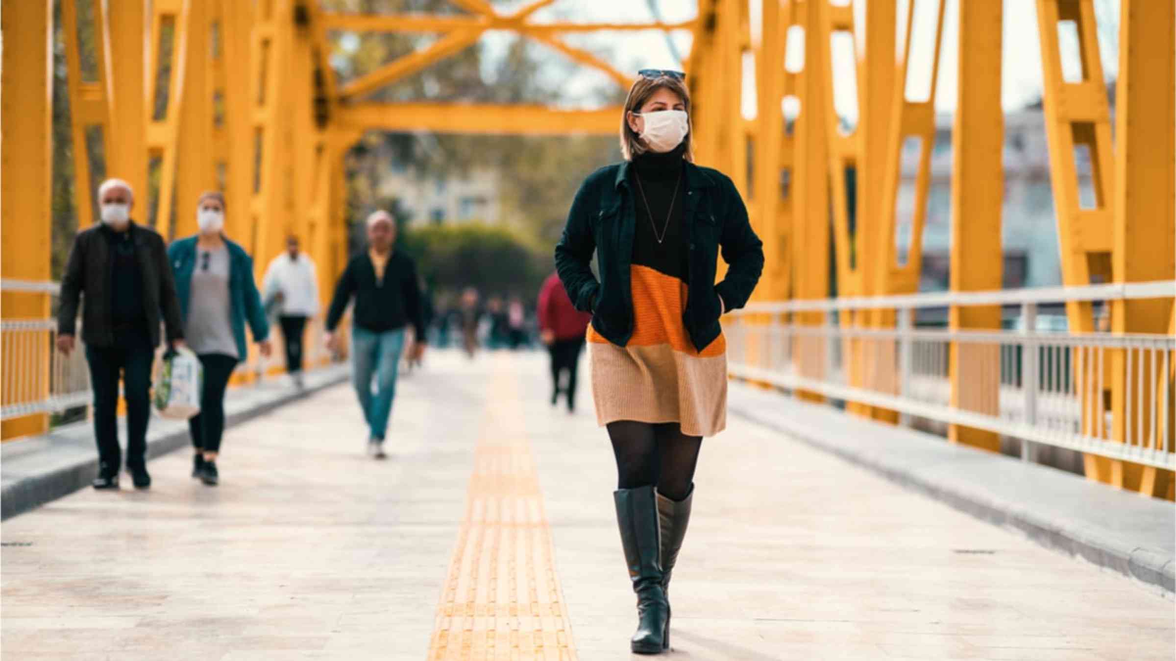 A woman wearing a face mask to protect herself from COVID-19 walking across a yellow bridge.